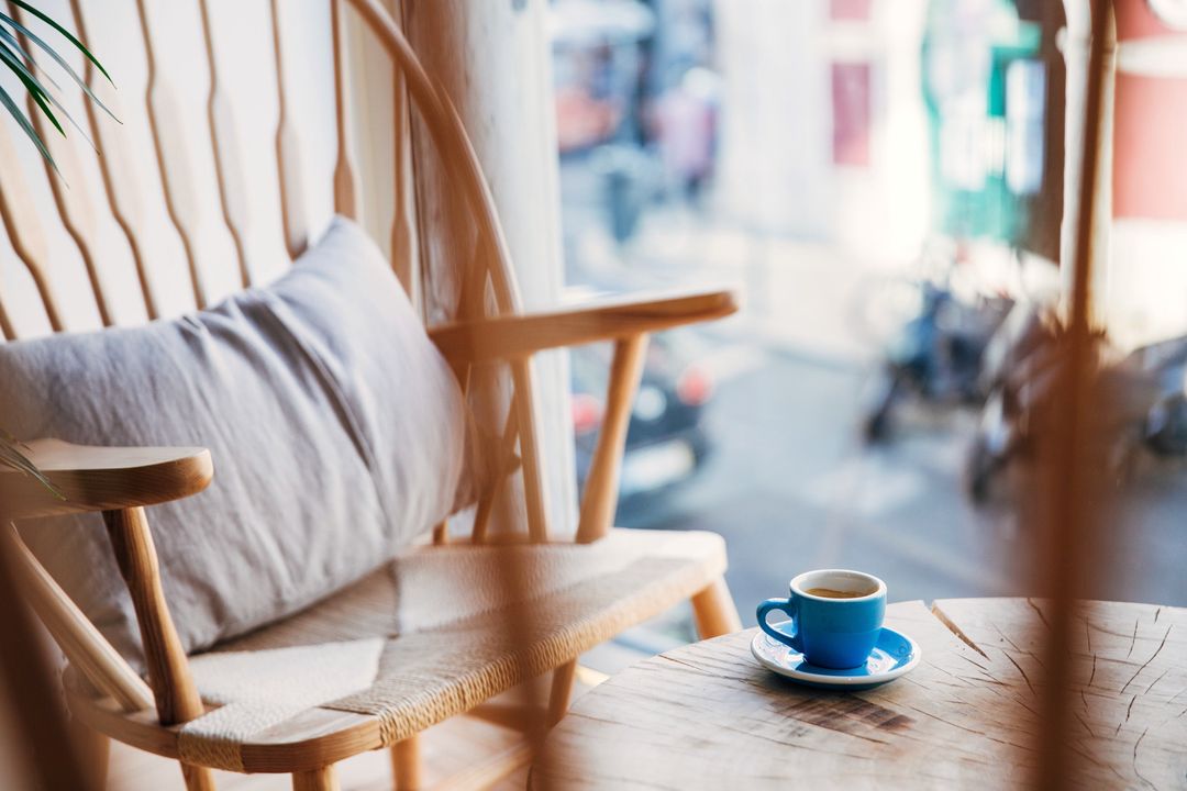 Ein Holzsessel mit Kissen am Fenster, auf dem kleinen Tisch eine blaue Espressotasse