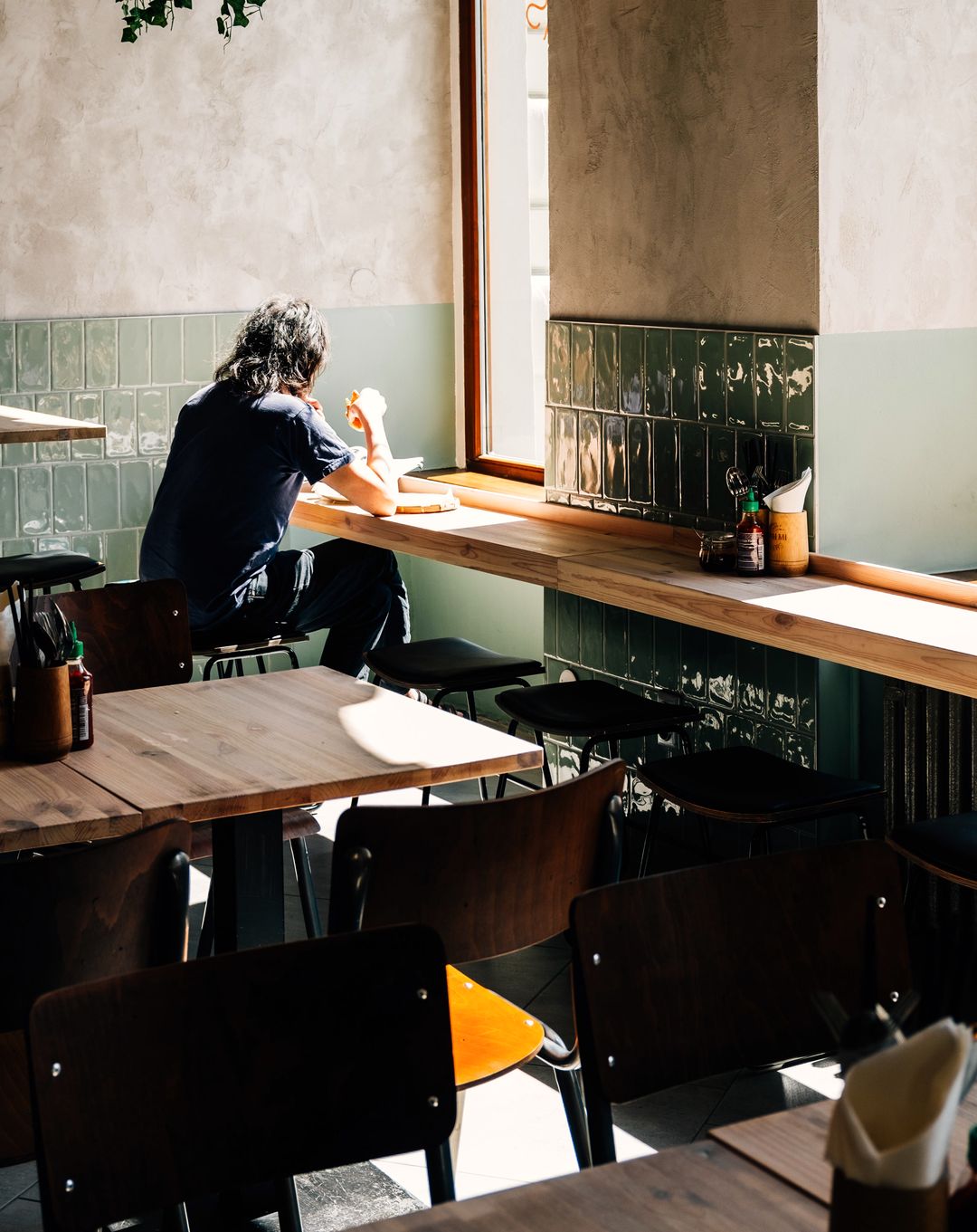 Ein Gast sitzt mit dem Rücken zum Raum an der Fensterbank eines sonnigen Cafés, davor Holztische und Stühle