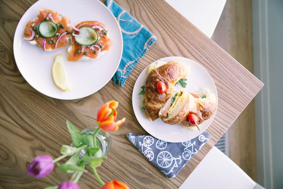 Von oben: ein Teller mit Lachs auf Brot, ein Croissant-Sandwich, Servietten und Tulpen auf einem Holztisch