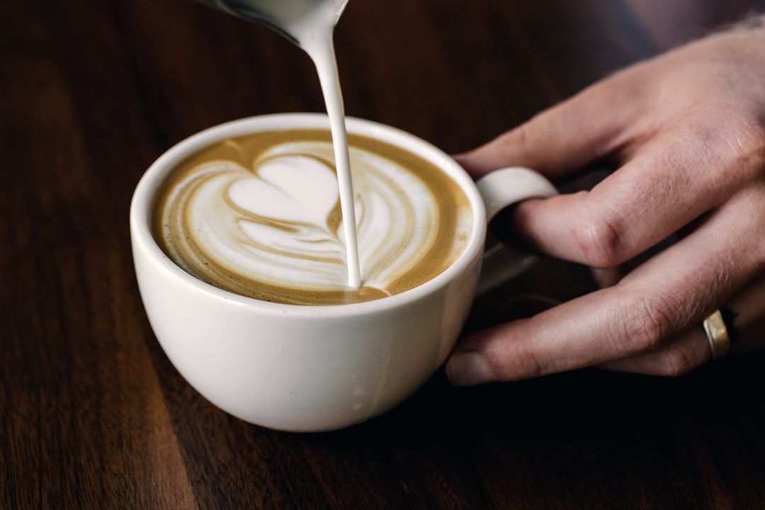 Milch wird in einen Cappuccino gegossen, eine Hand hält die Tasse