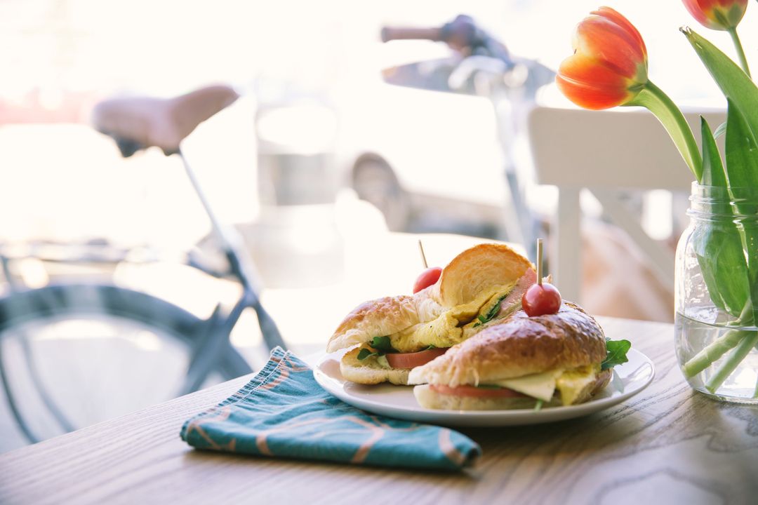 Ein Croissant-Sandwich mit Rührei und Tomate auf einem Teller am Fenster, daneben Tulpen
