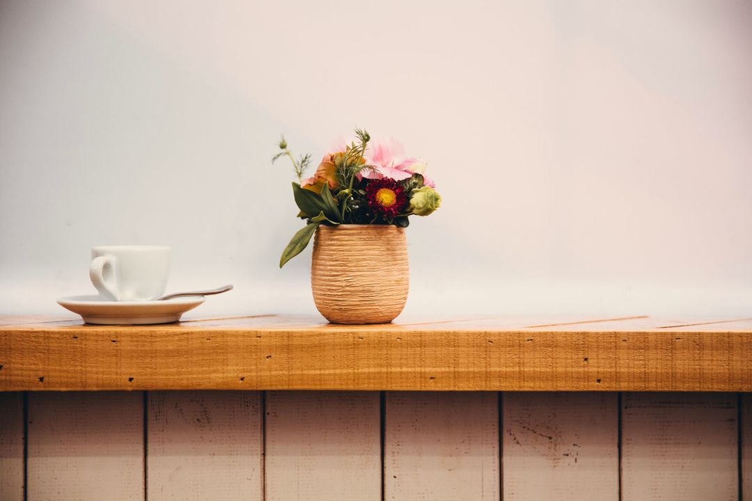 Eine Kaffeetasse und ein kleiner Blumenstrauß im Tontopf auf einer Fensterbank aus Holz vor einer sonnigen weißen Wand