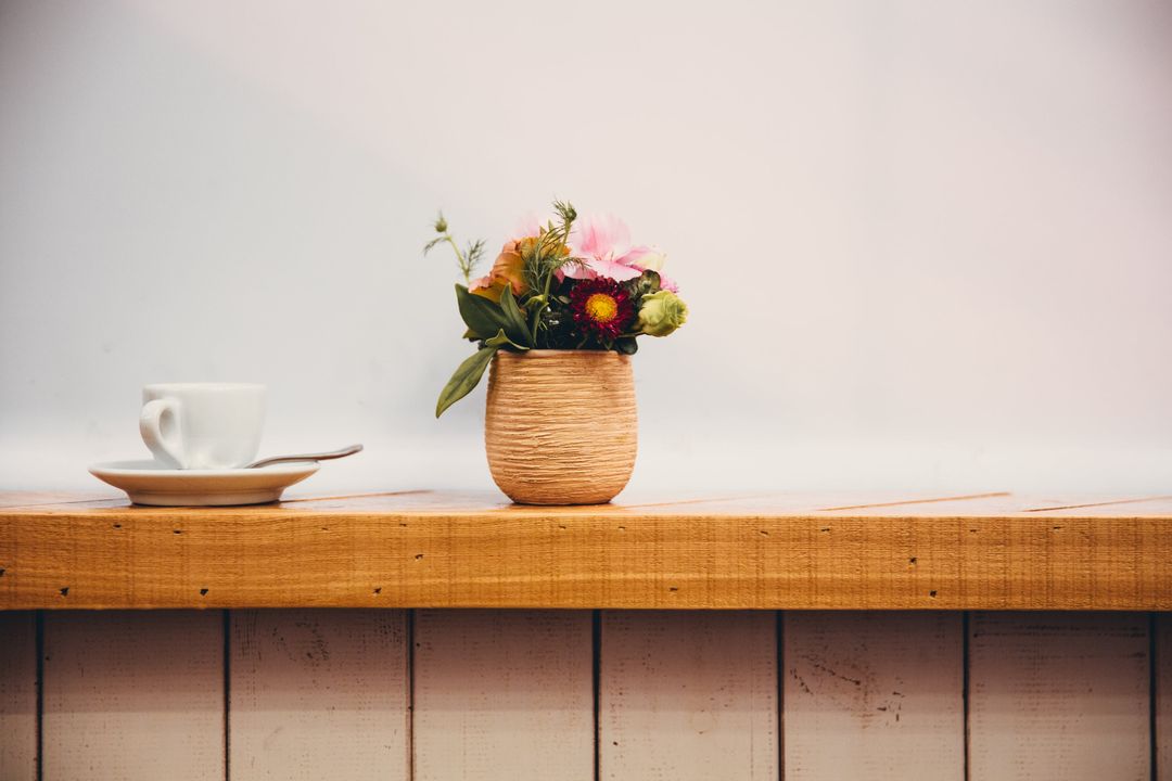 Eine Kaffeetasse und ein kleiner Blumenstrauß im Tontopf auf der Fensterbank vor einer weißen Wand