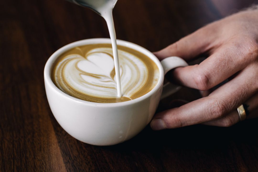 Milch wird in einen Cappuccino gegossen, eine Hand hält die Tasse