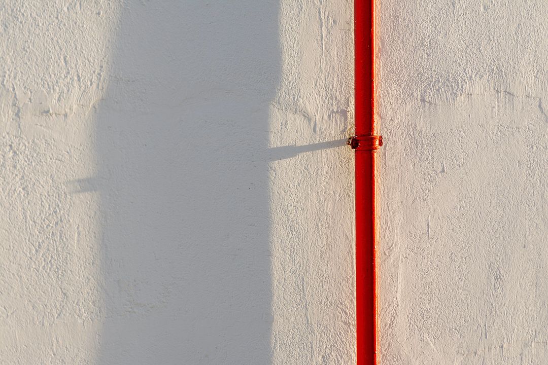 Ein rot lackiertes Heizungsrohr läuft senkrecht über eine sonnige weiße Wand, daneben der Schatten eines Hauses