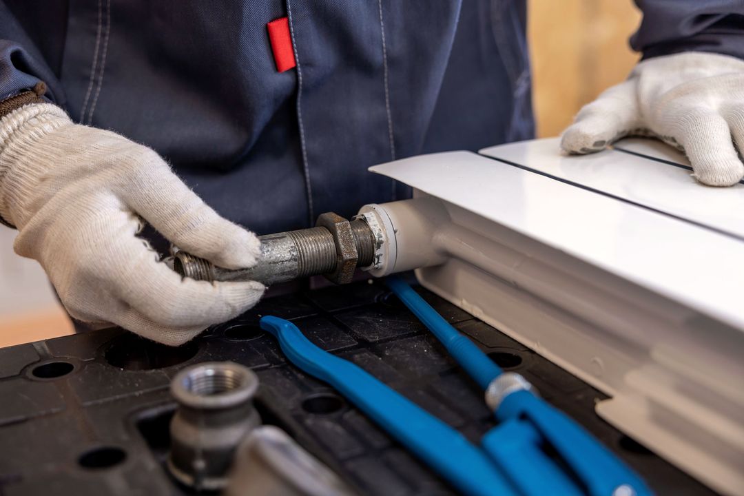 Hände in weißen Arbeitshandschuhen schrauben ein Anschlussstück an einen Heizkörper, daneben zwei blaue Rohrzangen