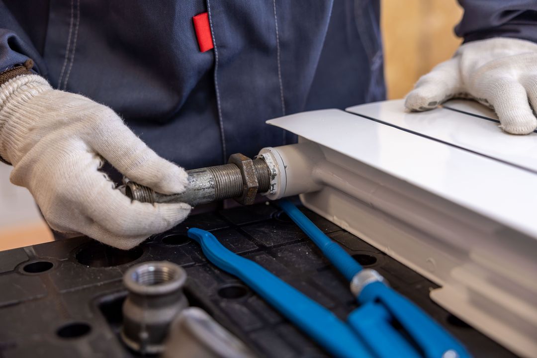 Hände in weißen Arbeitshandschuhen schrauben ein Anschlussstück an einen Heizkörper, daneben zwei blaue Rohrzangen
