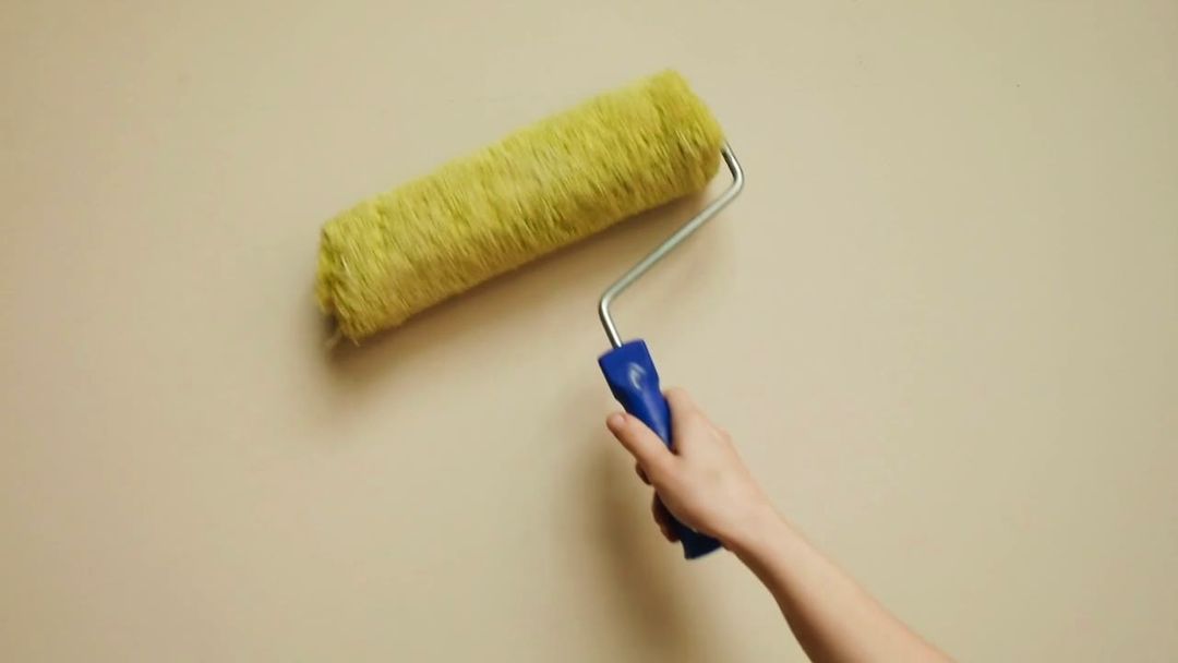 Eine gelbgrüne Farbrolle mit blauem Griff fährt über eine beige Wand