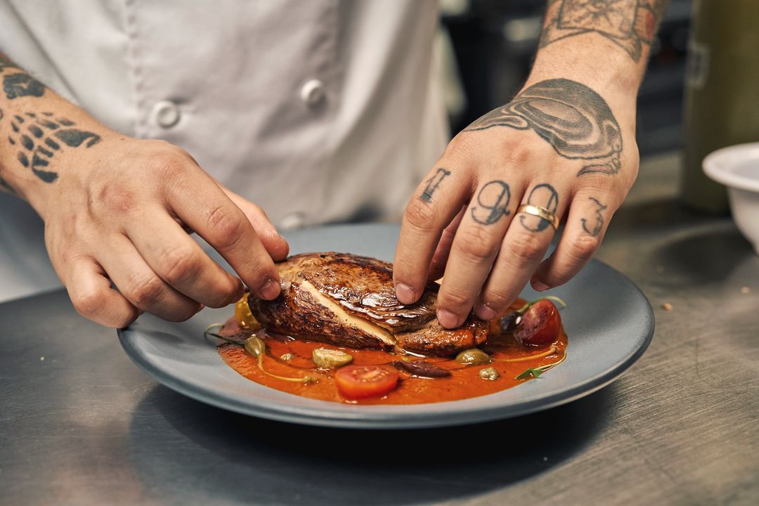 Zwei tätowierte Hände legen ein Stück Fisch auf eine Sauce aus Tomaten