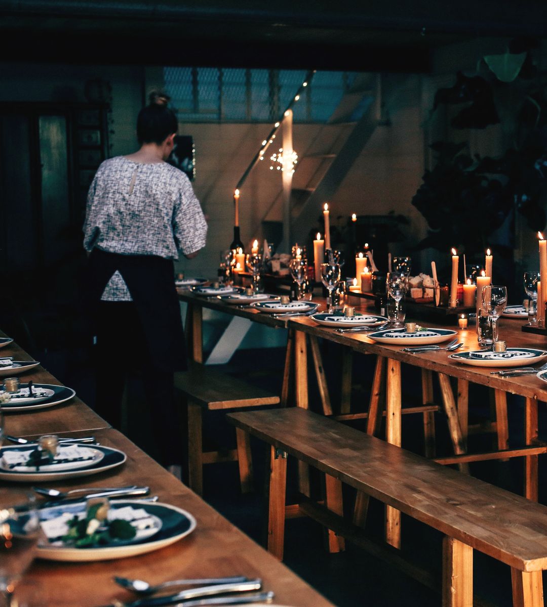 Eine lange Tafel mit Kerzen wird für den Abend eingedeckt, im Hintergrund eine Frau von hinten bei der Arbeit