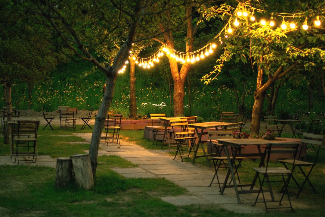 Tische und Stühle auf dem Rasen unter Obstbäumen, darüber Lichterketten in der Abenddämmerung