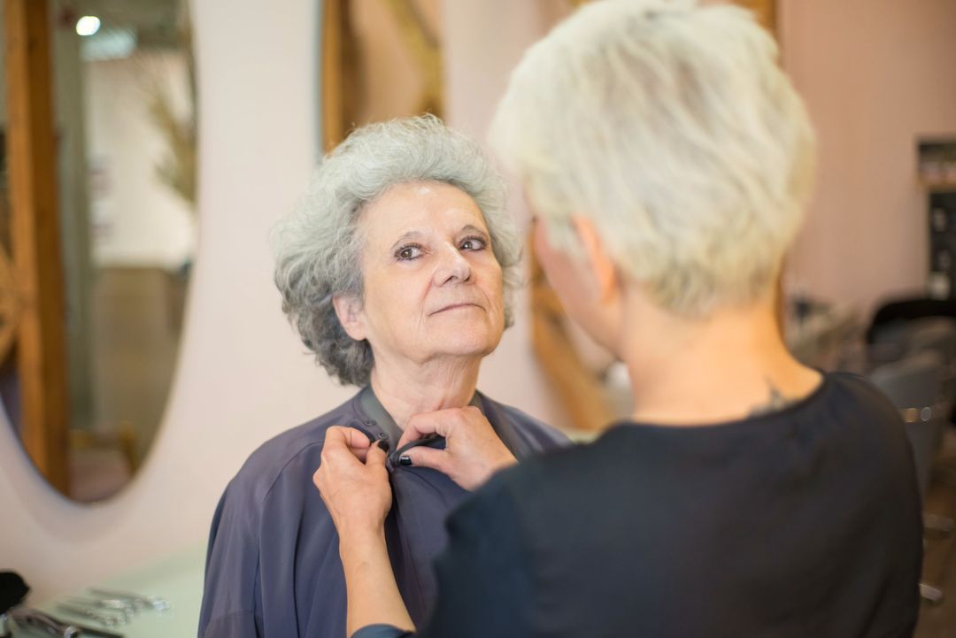 Die Friseurin legt einer Kundin mit grauem Haar den Umhang an, beide sehen sich an