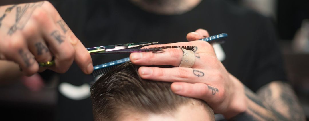 Ein Barbier mit tätowierten Händen schneidet mit Kamm und Schere das Deckhaar eines Herrenschnitts