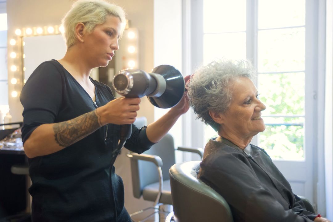 Die Friseurin föhnt einer lächelnden Kundin am hellen Fenster die Haare