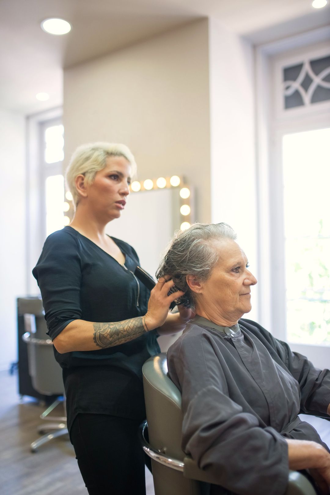 Eine Friseurin frisiert einer Kundin am hellen Fenster das graue Haar
