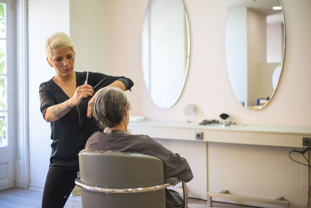 Eine Friseurin schneidet einer Kundin die Haare, an der hellen Wand hängen zwei ovale Spiegel, links fällt Licht durchs Fenster