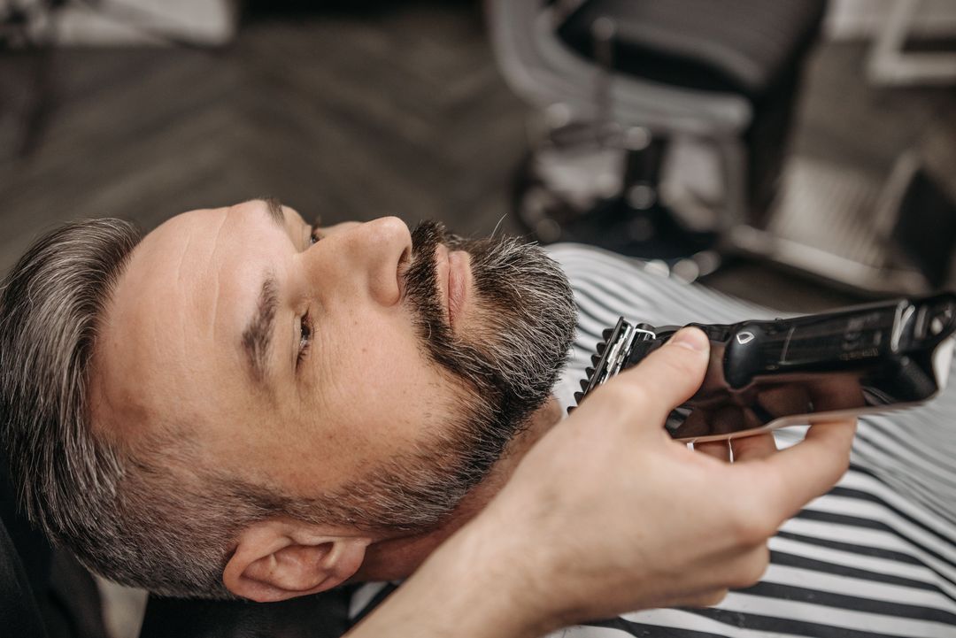 Ein Barbier trimmt einem Mann im zurückgelehnten Stuhl den grau melierten Bart mit der Maschine