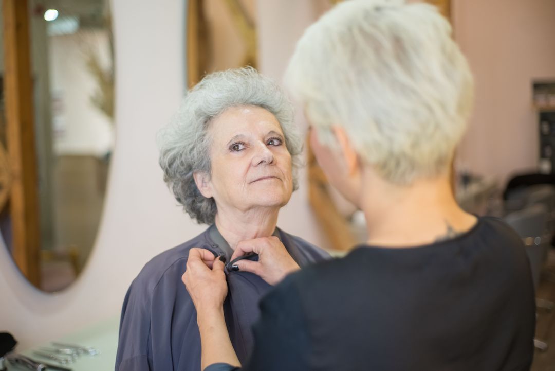 Die Friseurin legt einer Kundin mit grauem Haar den Umhang an, beide sehen sich an