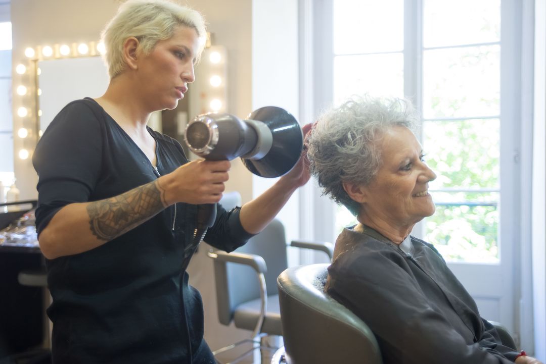 Die Friseurin föhnt einer lächelnden Kundin am hellen Fenster die Haare