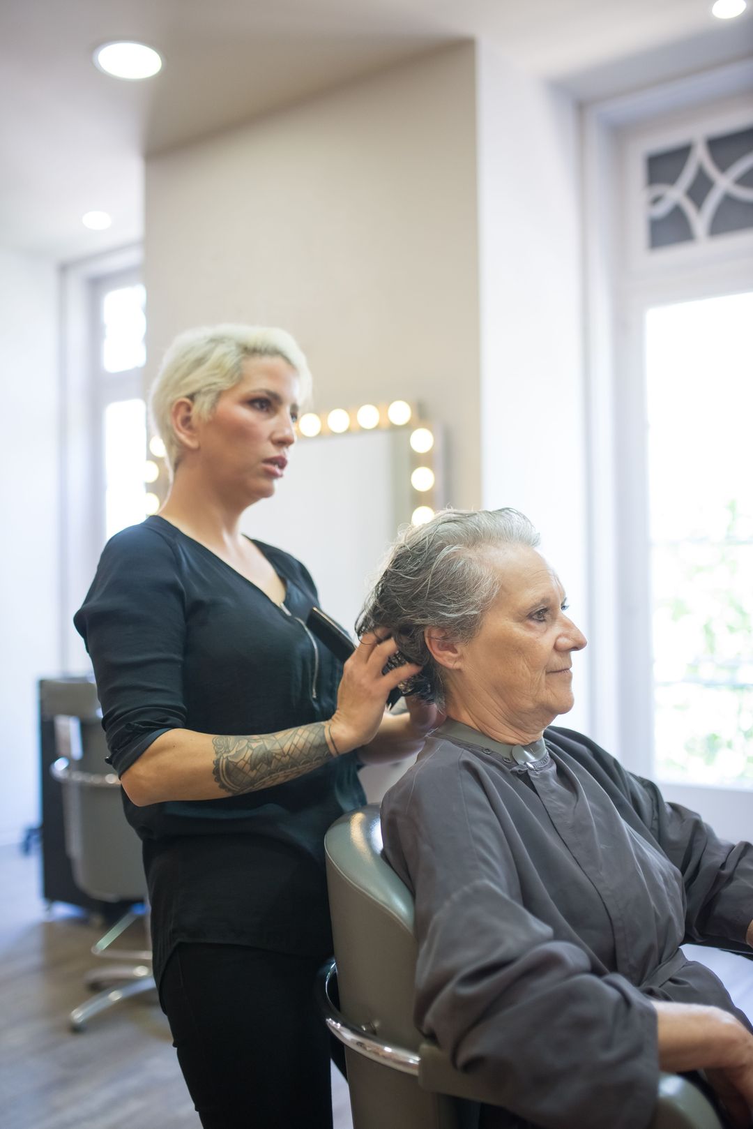 Eine Friseurin frisiert einer Kundin am hellen Fenster das graue Haar