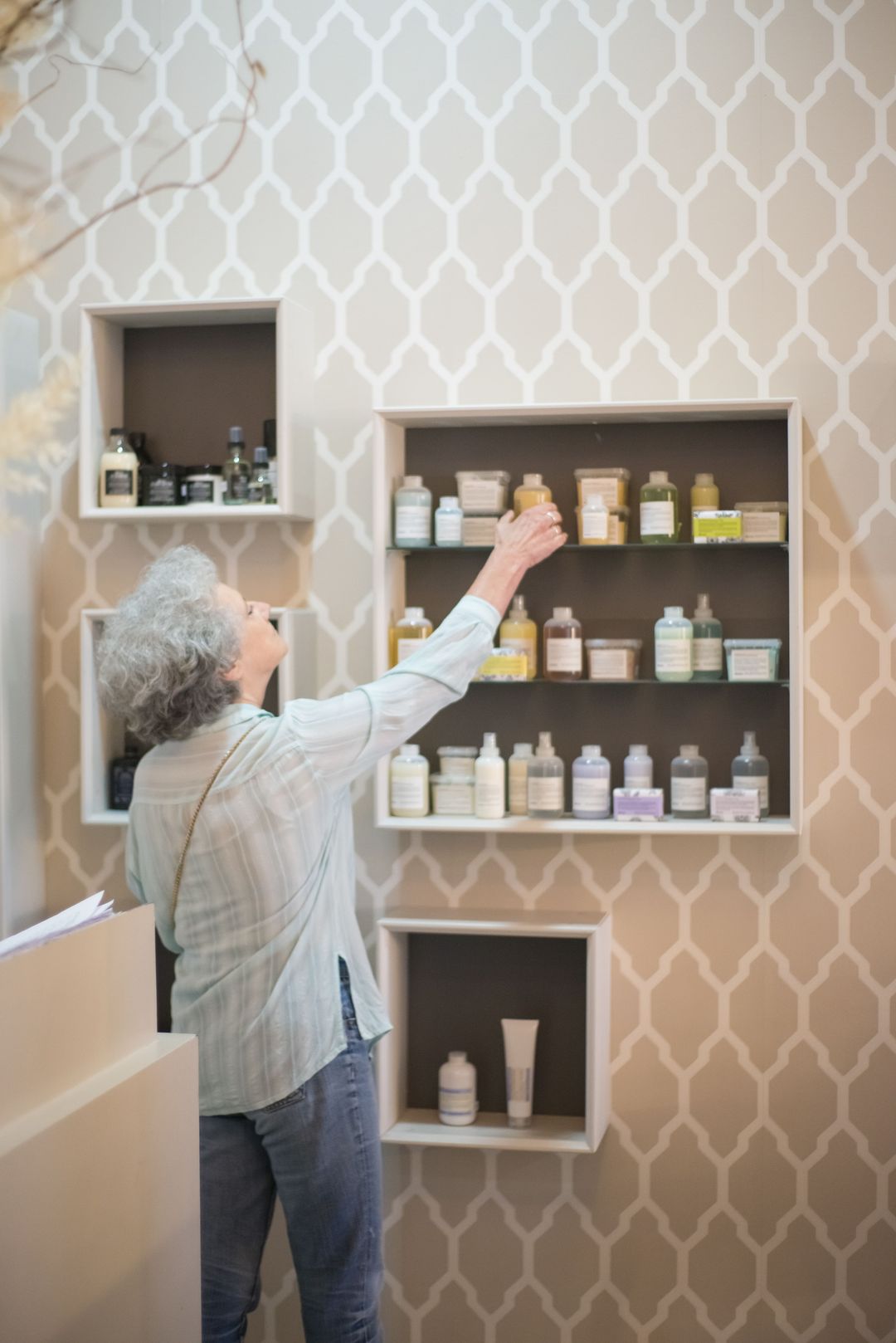 Eine Kundin greift in das Wandregal mit Shampoos und Pflegeprodukten vor einer gemusterten Tapete