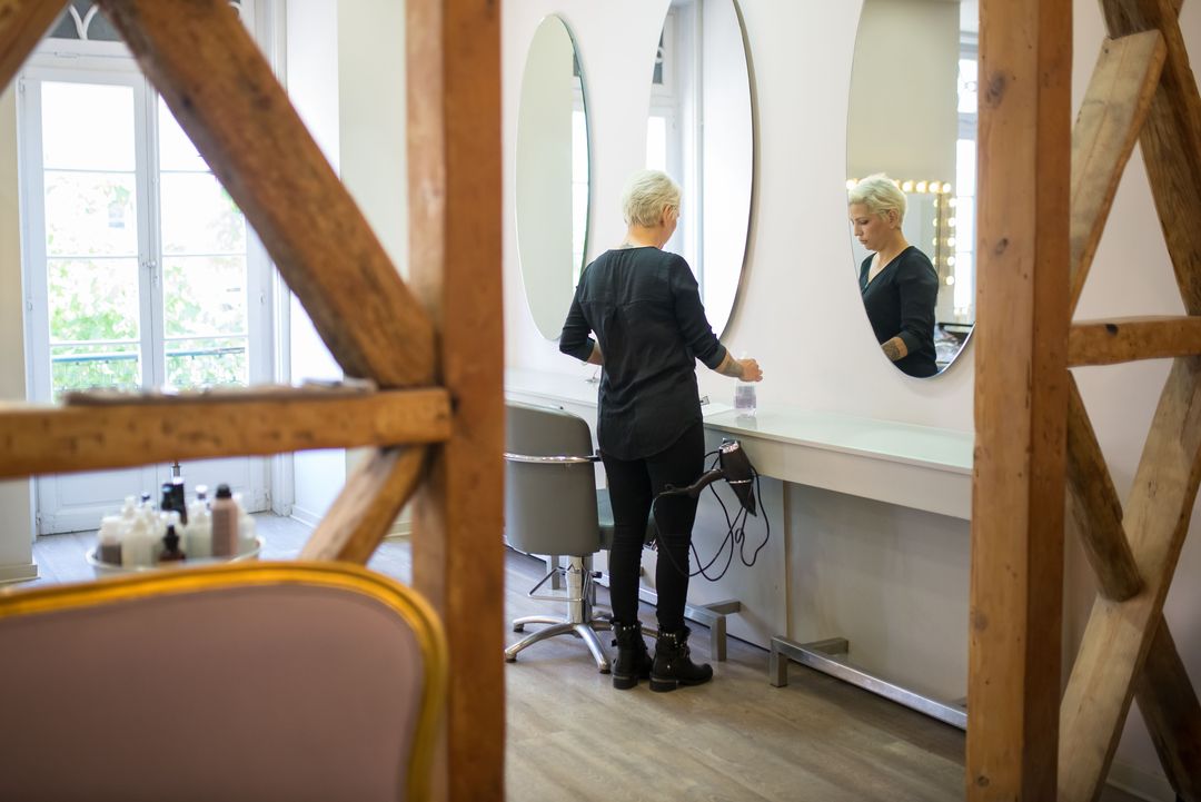 Durch die Balken des Fachwerks sieht man eine Friseurin an ihrem Platz vor einem ovalen Spiegel, links ein hohes Fenster