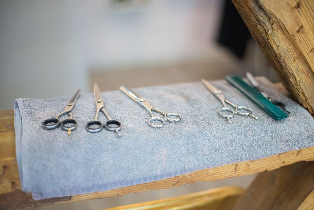 Fünf Scheren und ein Kamm liegen auf einem grauen Handtuch über einem Holzbalken, dahinter der helle Salon