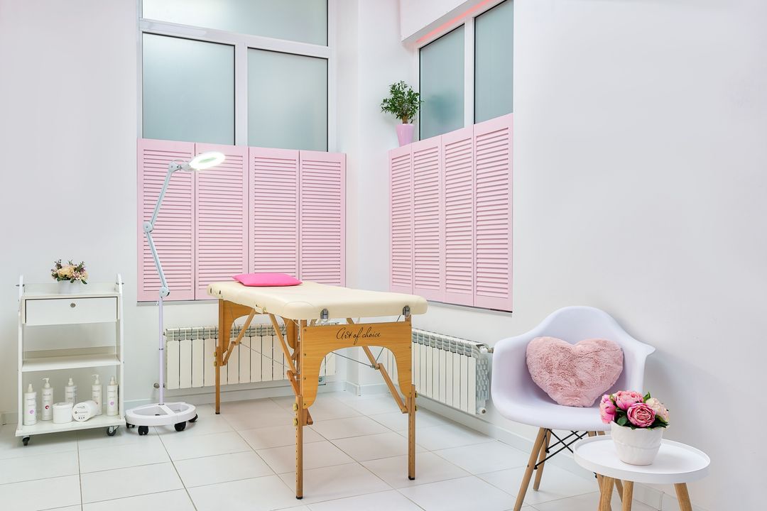 Der helle Behandlungsraum von der Seite: Liege, Lupenleuchte, ein Wagen mit Pflegeprodukten und Blumen