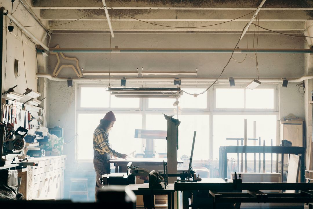 Ein Tischler steht im Gegenlicht an einem Arbeitstisch in der Werkstatt zwischen Werkzeug und Maschinen