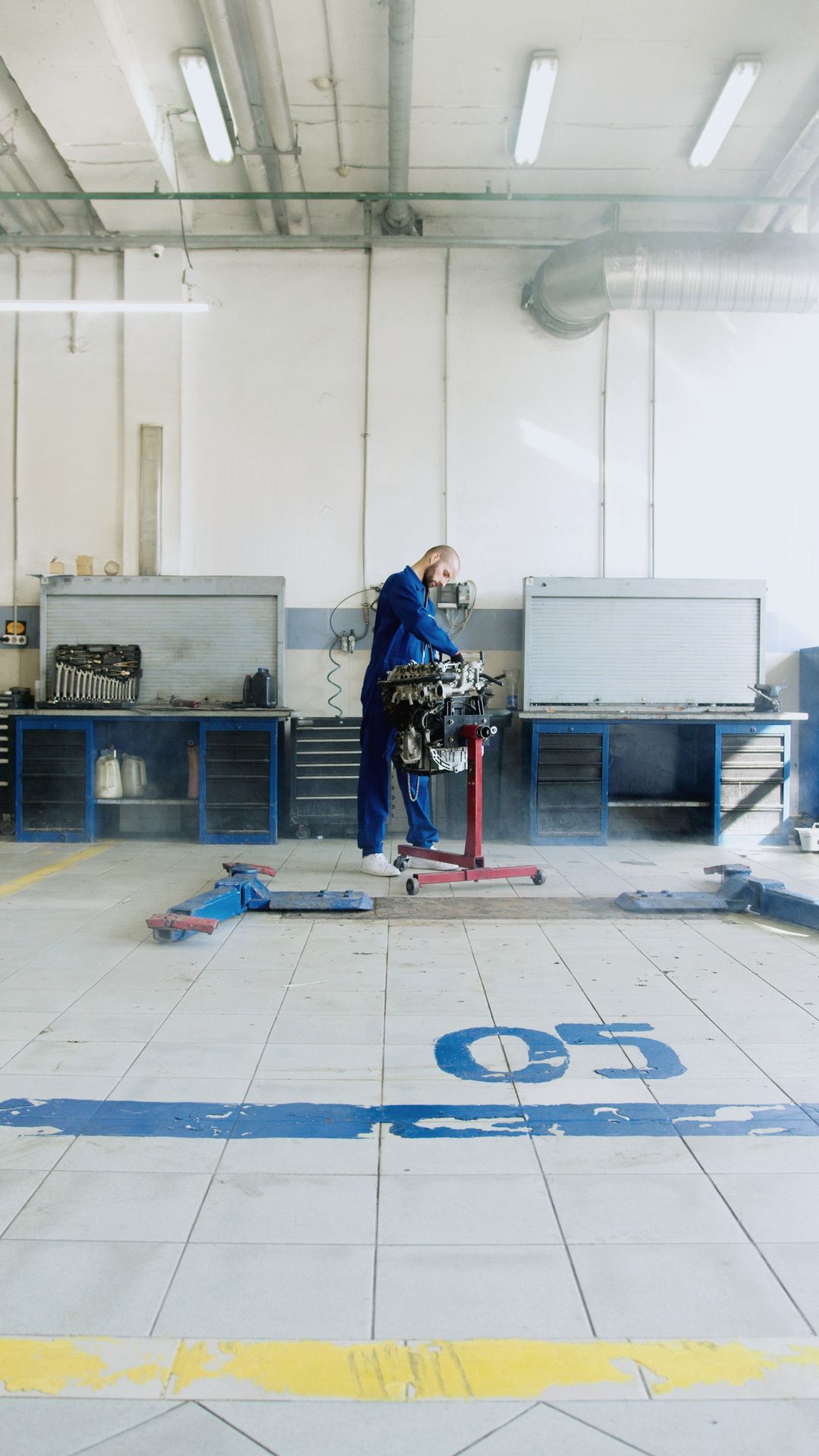 Die helle Halle mit Werkbänken, ein Mechaniker arbeitet an einem Motor auf dem Montageständer, auf dem Boden die Nummer 05