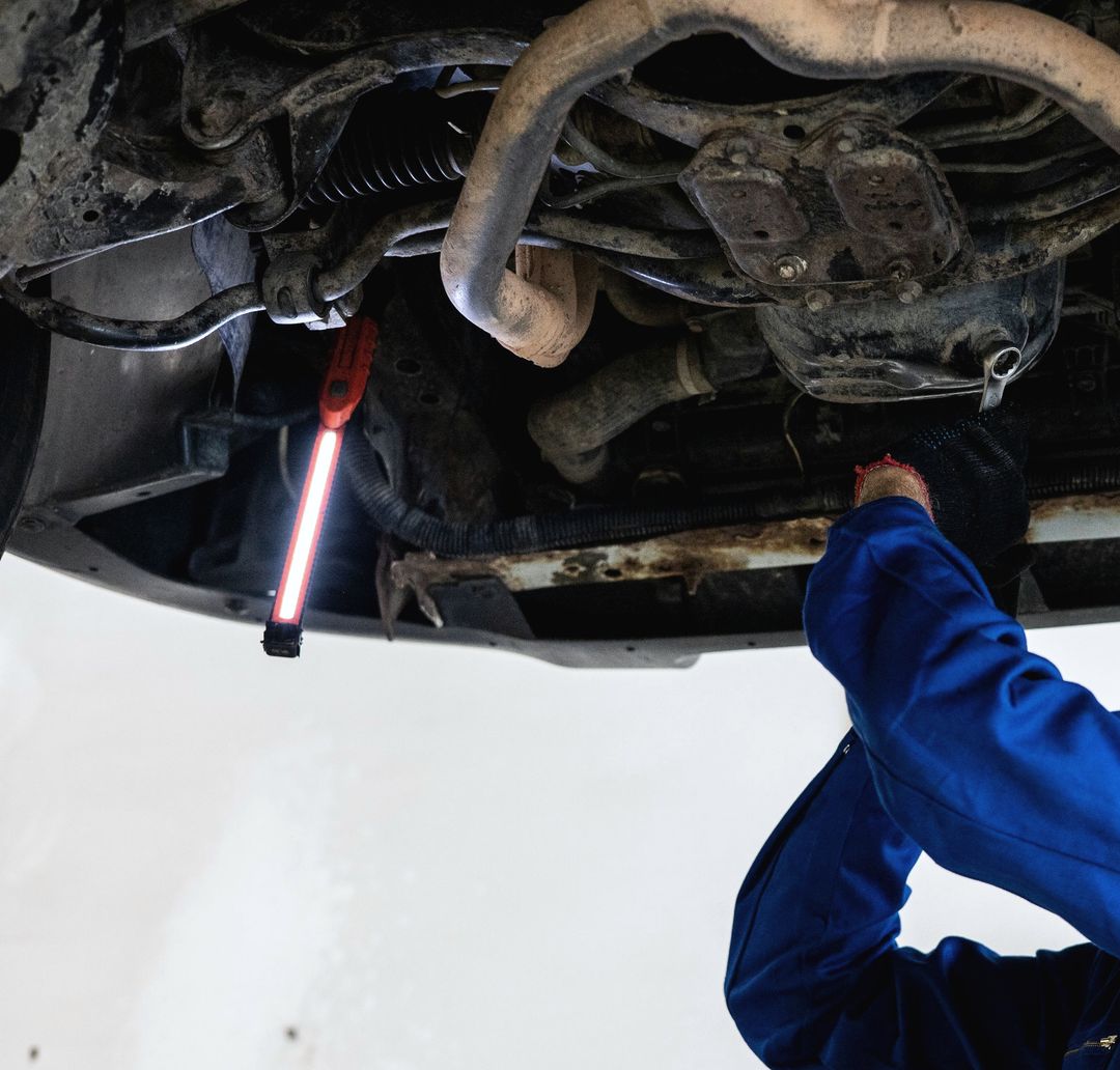 Ein Mechaniker im blauen Overall leuchtet mit einer Handlampe den Unterboden eines Autos auf der Hebebühne ab