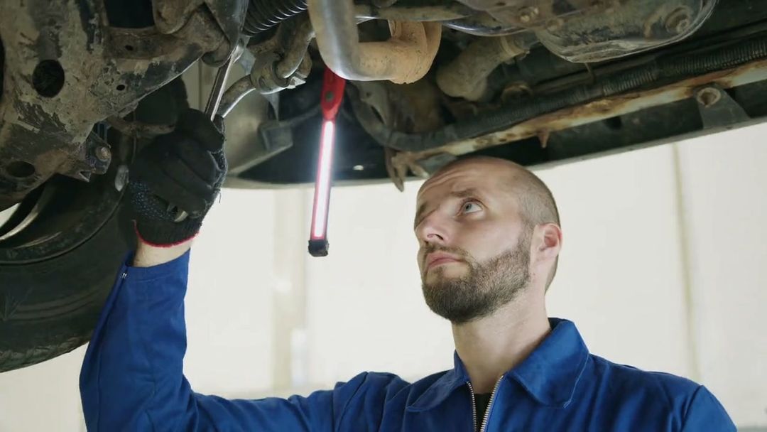 Ein Mechaniker im blauen Overall schaut mit einer Handlampe nach oben auf den Unterboden eines angehobenen Autos