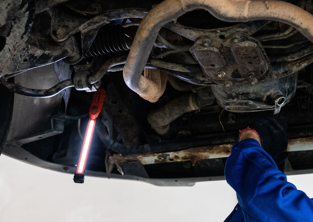 Ein Mechaniker im blauen Overall leuchtet mit einer Handlampe den Unterboden eines Autos auf der Hebebühne aus